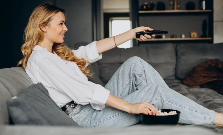 Eine blonde Frau auf einem grauen Sofa, die eine Hand in eine Schüssel voll Popcorn hält und die andere mit einer Fernbedienung in der Hand nach vorne streckt